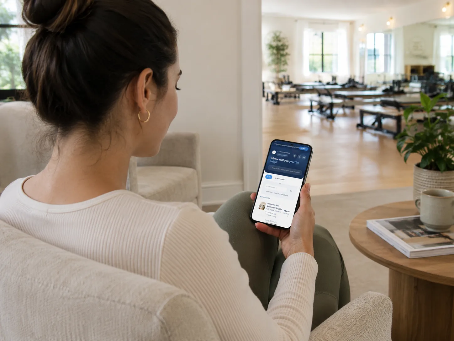 A practitioner sitting with her phone, choosing a room, with a reformer studio behind her.